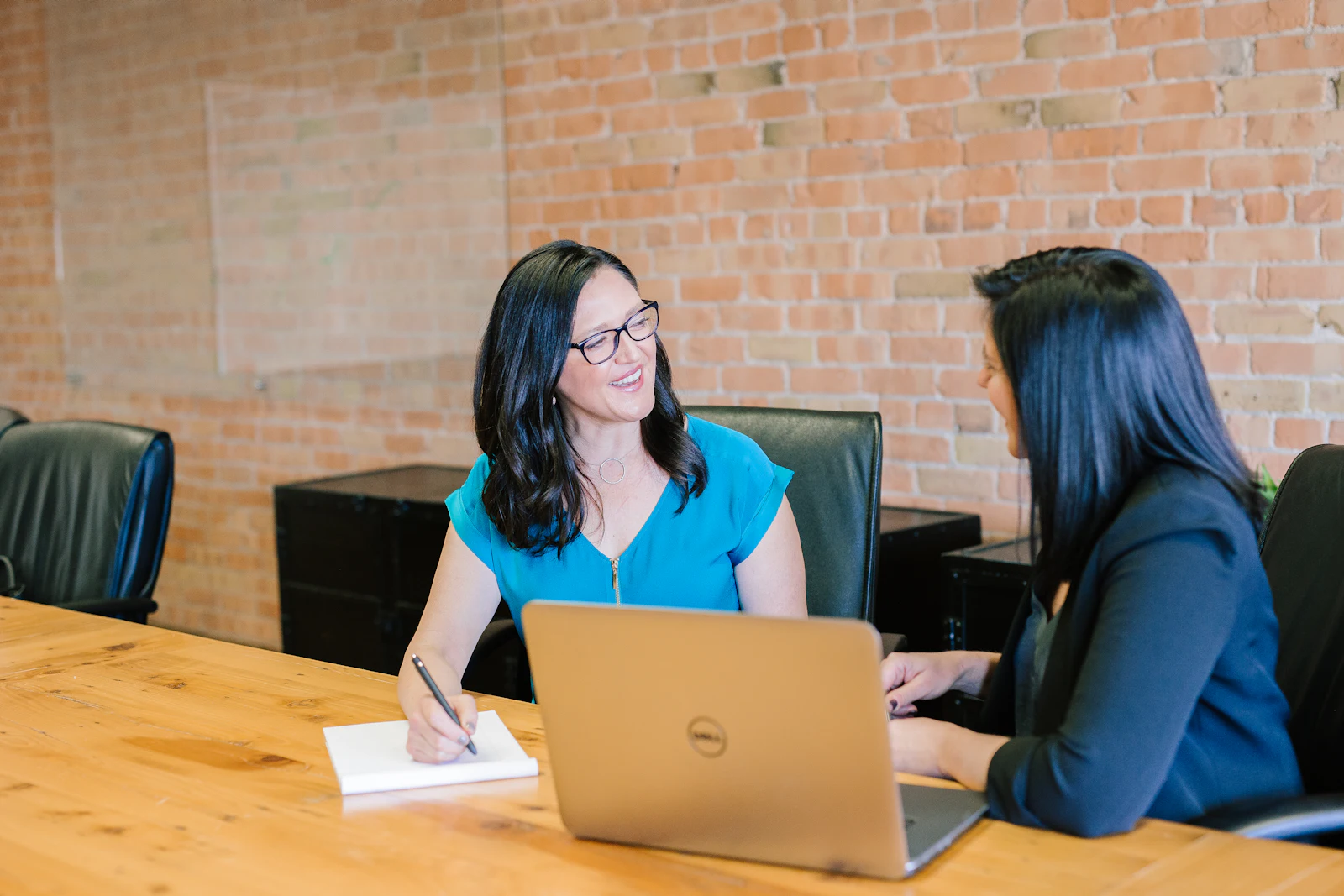 Two people talking through a project at a meeting table, one taking notes beside a laptop.